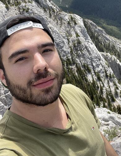 A selfie of a man stands on a mountain wearing a green shirt and baseball hat