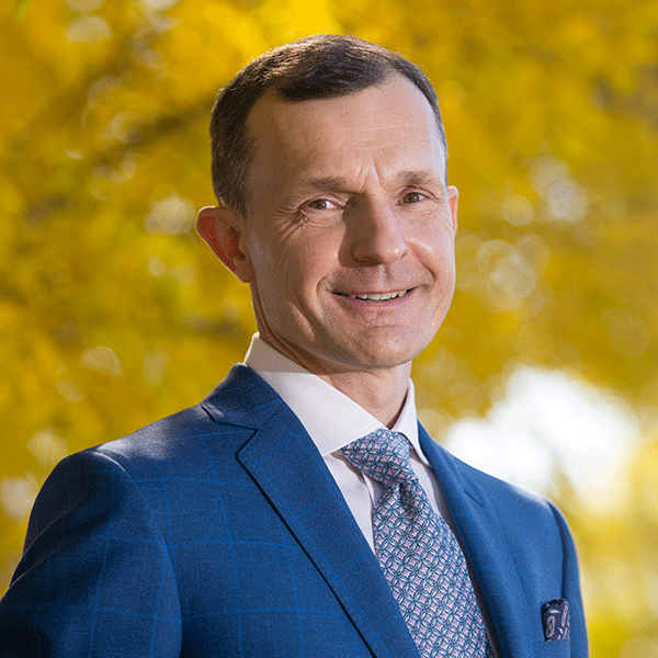 Tom Stelfox is wearing a blue suit and is smiling. He is standing outside in front of an autumnal tree. 