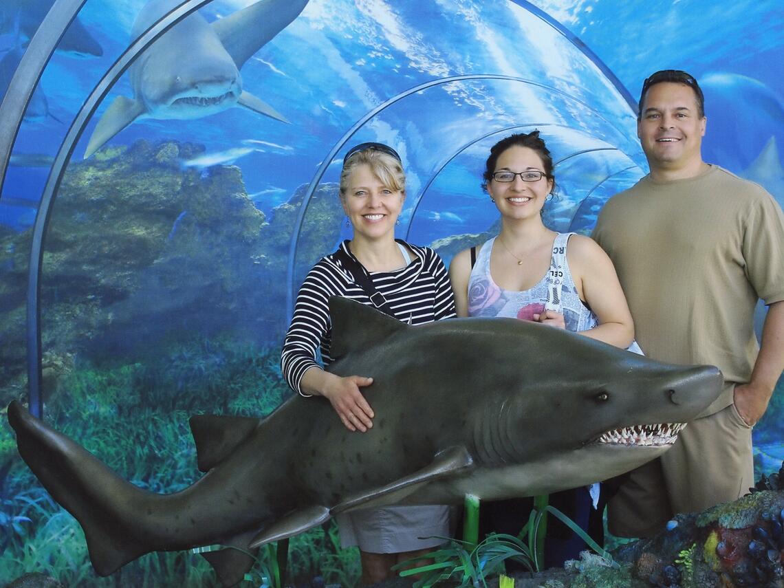 three people pose with a life-size model of a shark, against an aquarium background