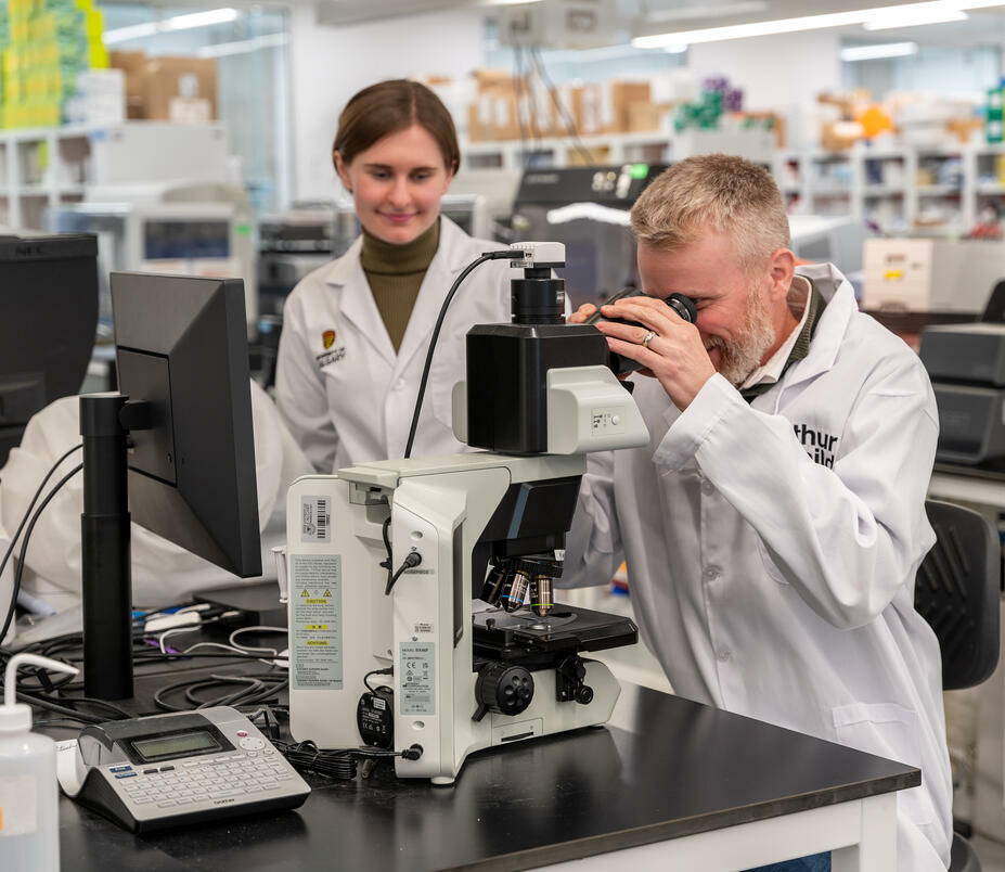 man in a lab coat looks into a microscope while a woman in a lab coat looks on
