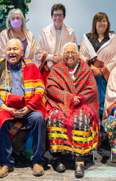 A group of people wearing ribbon skirts and blankets smile at the camera